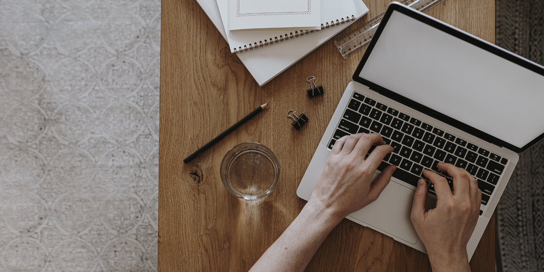 Женски ръце пишат на лаптоп на дървено бюро, до чаша вода и тефтери, момент на работа и нужда от пауза.