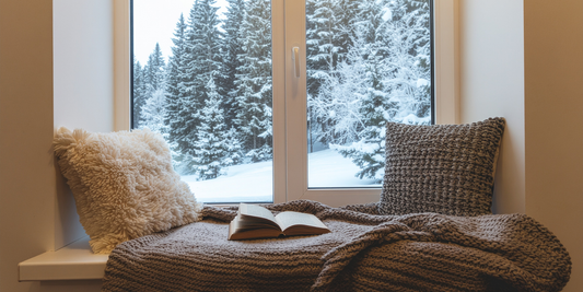 Тиха зимна сцена у дома с място за сядане до прозорец, книга и мека светлина, гледаща към заснежена гора