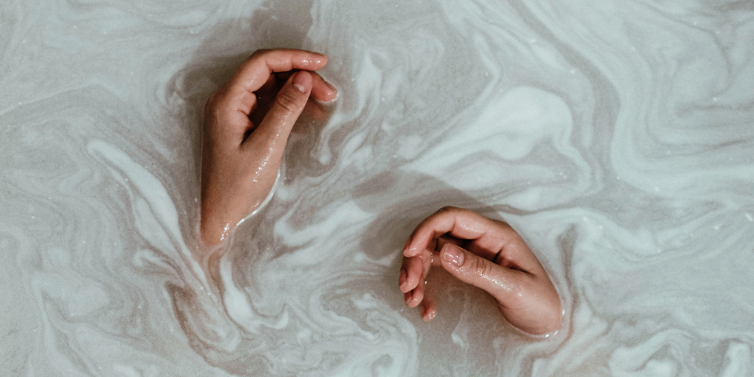 Женски ръце, потопени в топла мътна вода, създаваща усещане за релакс и self-care ритуал.