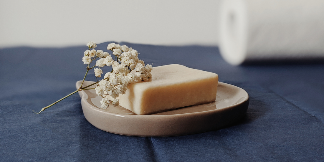 Натурален сапун върху керамична сапунерка, поставен на тъмносиня ленена повърхност с малък сух цвят до него.