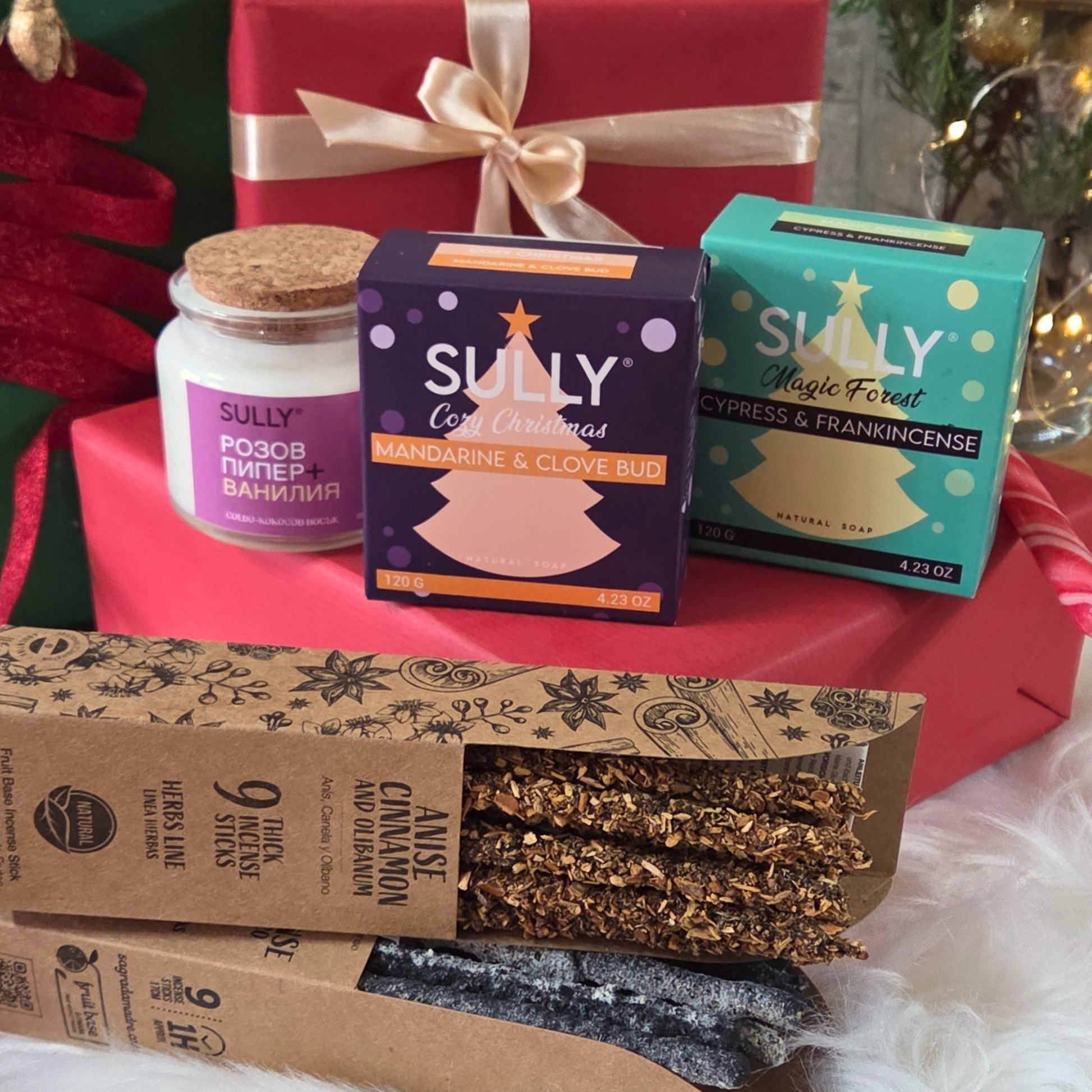 Празничен комплект „Коледен аромат & светлина“ SULLY Naturals – свещ, сапуни и ароматни пръчици, подредени сред коледни подаръци.