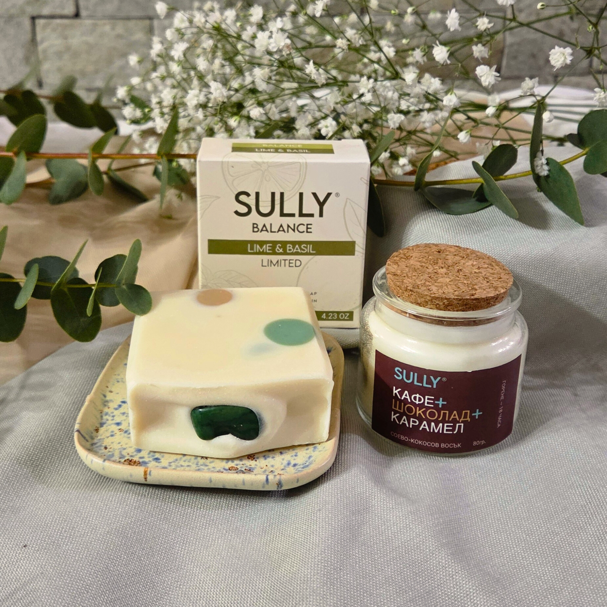 Комплект „Душ и аромат“ с кристален сапун BALANCE (лайм и босилек), керамична сапунерка и соева свещ „Кафе, шоколад и карамел“, заснет на текстил с евкалипт и цветя на фон.