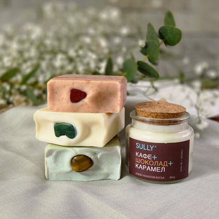 Трите кристални сапуна SULLY Naturals FOCUS, BALANCE и IMPULSE, подредени един върху друг до соева ароматна свещ „Кафе, шоколад и карамел“ на нежен фон с цветя