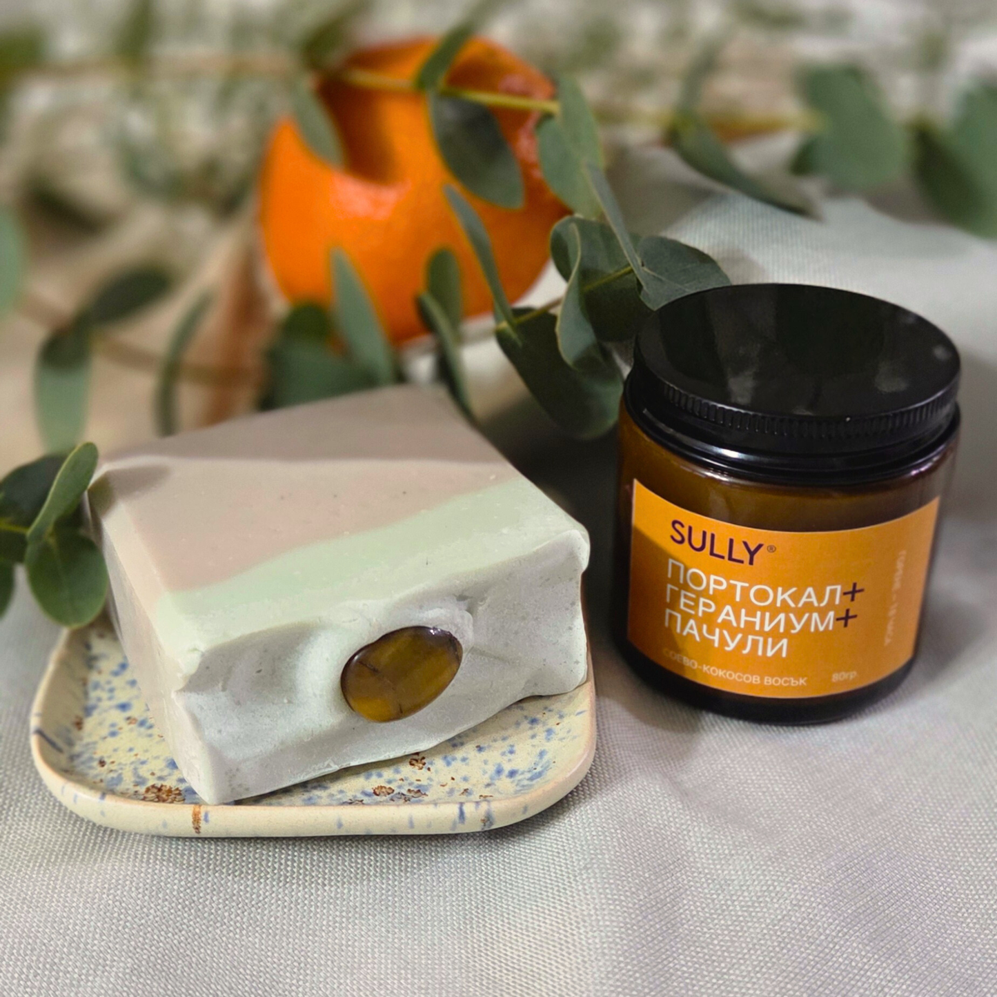 Комплект „Душ и аромат“ с кристален сапун FOCUS (зелена глина и пачули), керамична сапунерка и соева свещ „Портокал, гераниум и пачули“, заснет на текстил с евкалипт и портокал на фон.