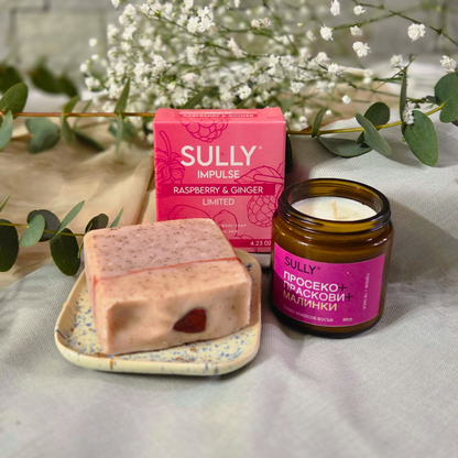 Комплект „Душ и аромат“ с кристален сапун IMPULSE (малина и джинджифил), керамична сапунерка и соева свещ „Просеко, праскови и малинки“, заснет на текстил с евкалипт и цветя на фон.