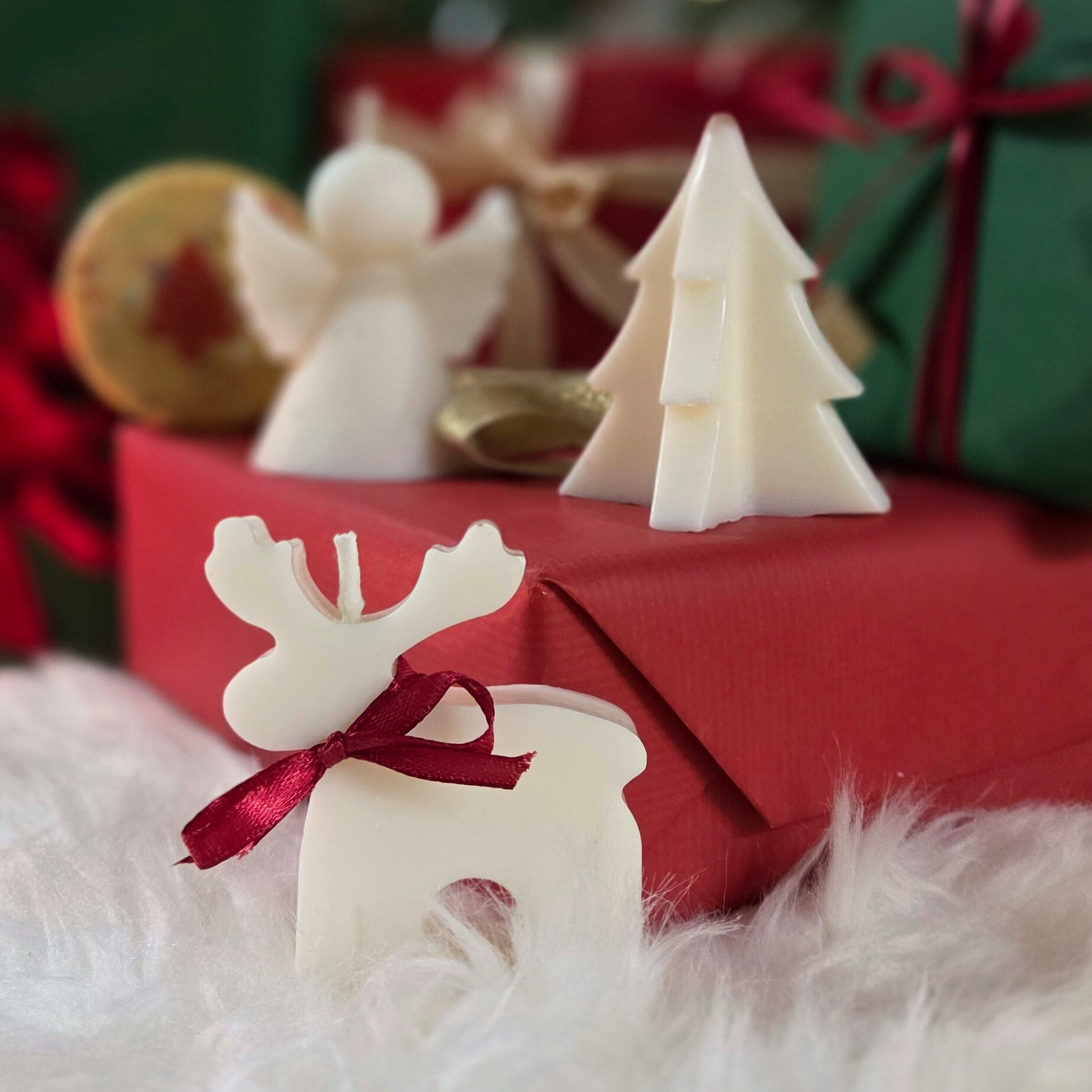 Коледна свещ във форма на еленче с червена панделка, поставена върху подаръци и бял пухкав фон.