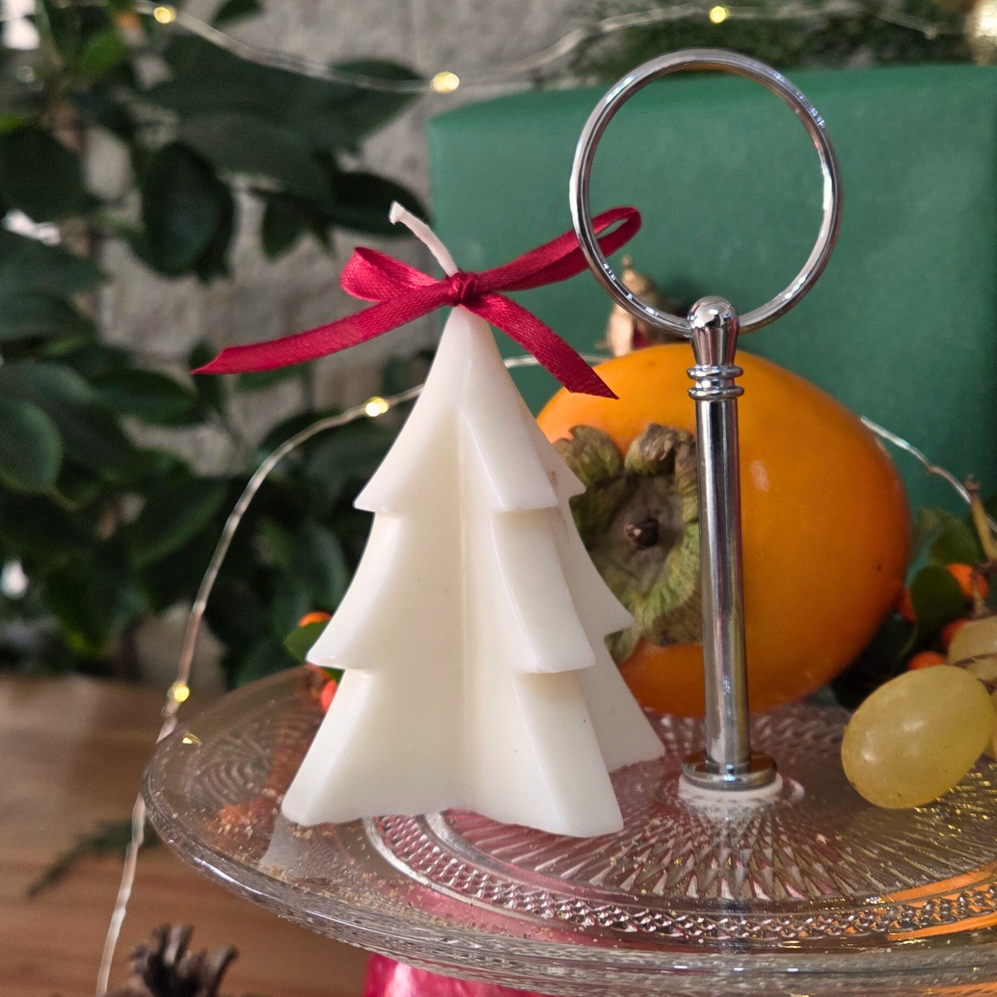 Коледна свещ във форма на елхичка с червена панделка, поставена върху стъклена поставка с празничен фон.