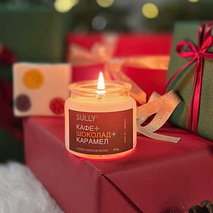 Ароматна свещ „Кафе, шоколад и карамел“ SULLY Naturals, поставена върху коледни подаръци с топла светлина.
