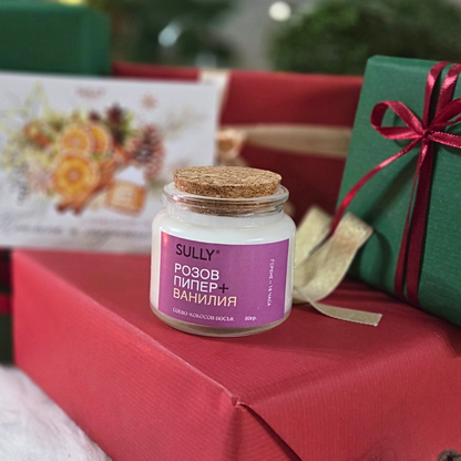 Ароматна свещ „Розов пипер и ванилия“ SULLY Naturals, заснета върху подаръци и коледна картичка.