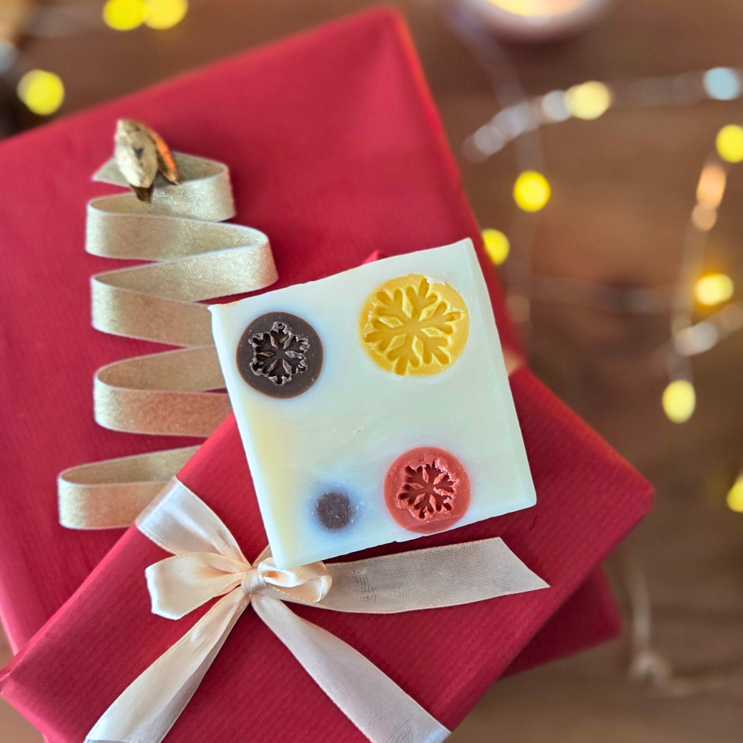 Ръчно изработен сапун „Уютна Коледа“ с цветни снежинки, поставен върху червен подарък с панделка и златна декорация.