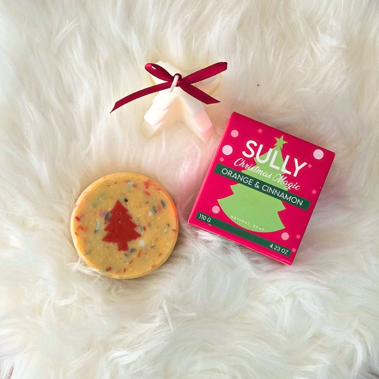 Сапун „Вълшебна Коледа“ SULLY Naturals с коледна опаковка, поставен върху бял пухкав фон до свещ във форма на елха.
