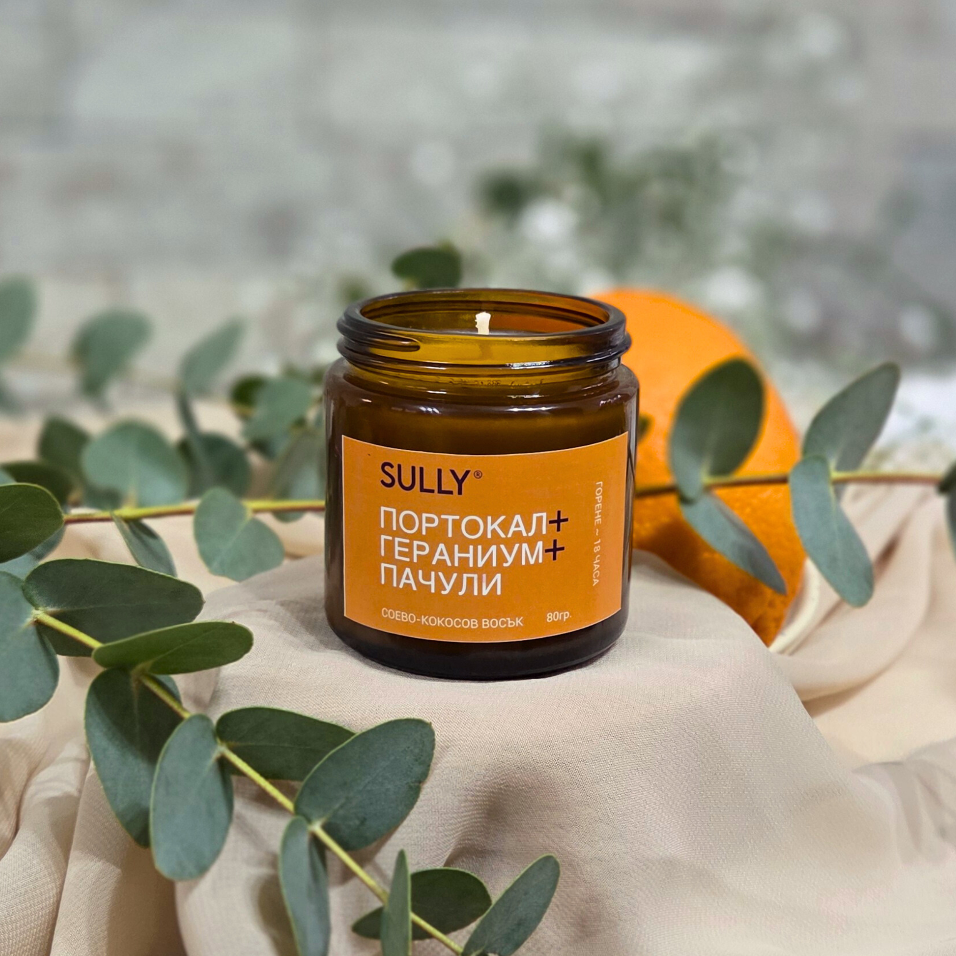 Соева ароматна свещ SULLY „Портокал, гераниум и пачули“ 80 г с портокал и евкалипт върху нежен плат на светъл фон
