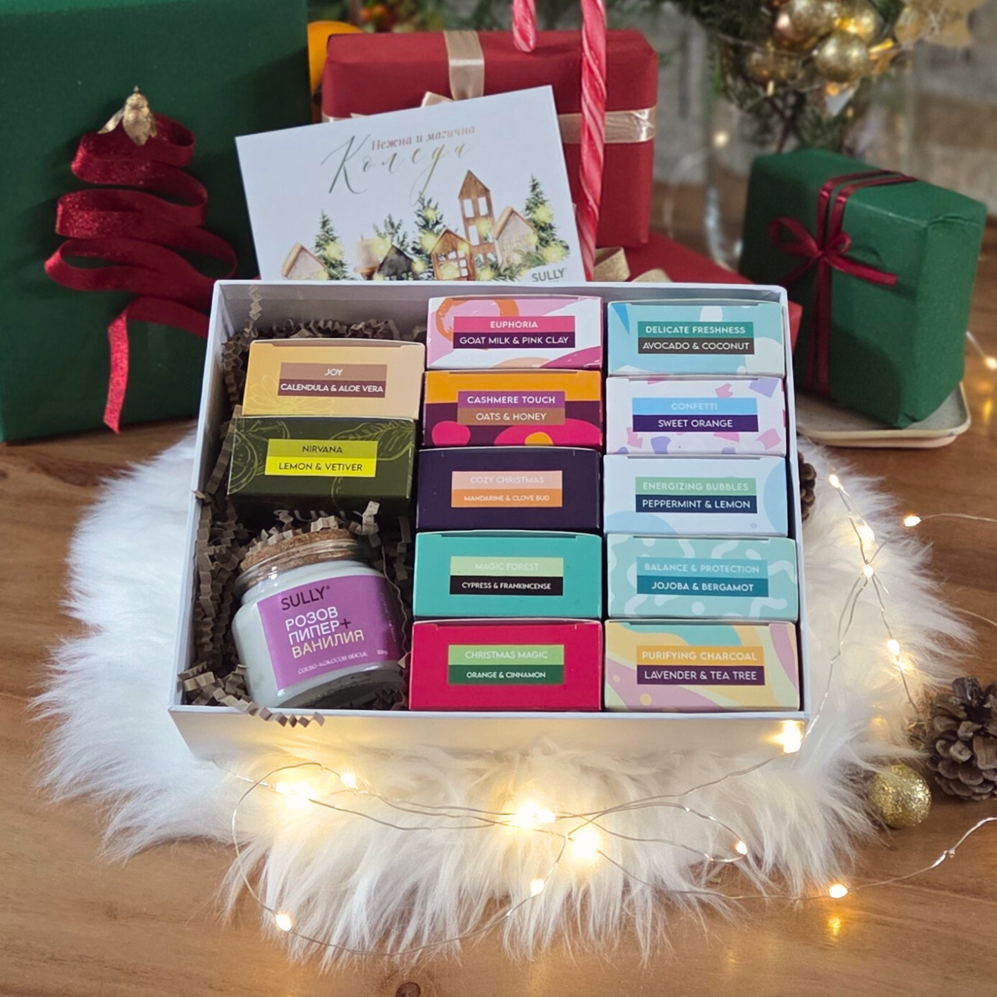 одаръчен комплект с 12 натурални сапуна SULLY в цветни опаковки, свещ и празнична картичка, подредени върху пухкав бял фон с лампички.