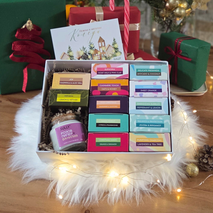 одаръчен комплект с 12 натурални сапуна SULLY в цветни опаковки, свещ и празнична картичка, подредени върху пухкав бял фон с лампички.