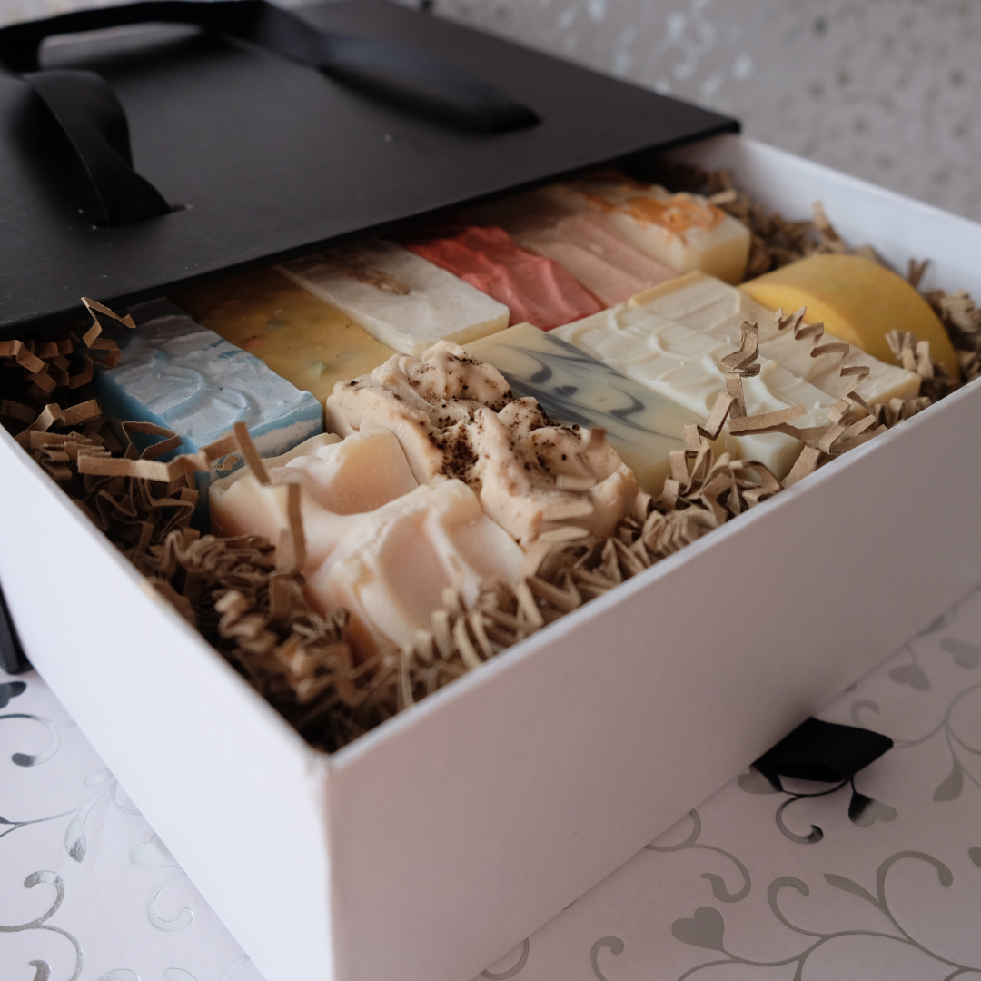 Подаръчна кутия с натурални сапуни SULLY, подредени в бяла кутия с черен капак, всеки с различен цвят и текстура, върху декоративна хартия.