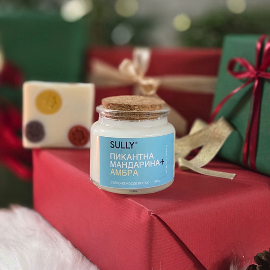 Ароматна свещ „Пикантна мандарина и амбър“ SULLY Naturals, поставена сред празнична украса в червено и златно