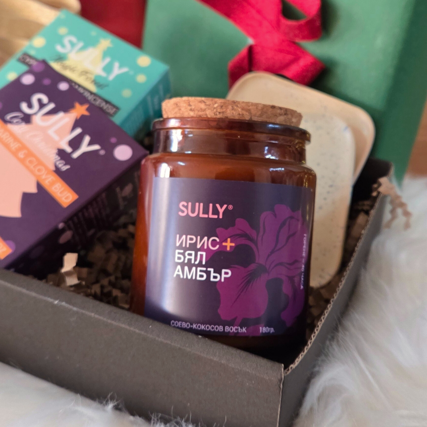 Свещ „Ирис и бял амбър“ от комплекта „Зимна грижа“ SULLY Naturals, заснета сред празнична украса и сапуни.