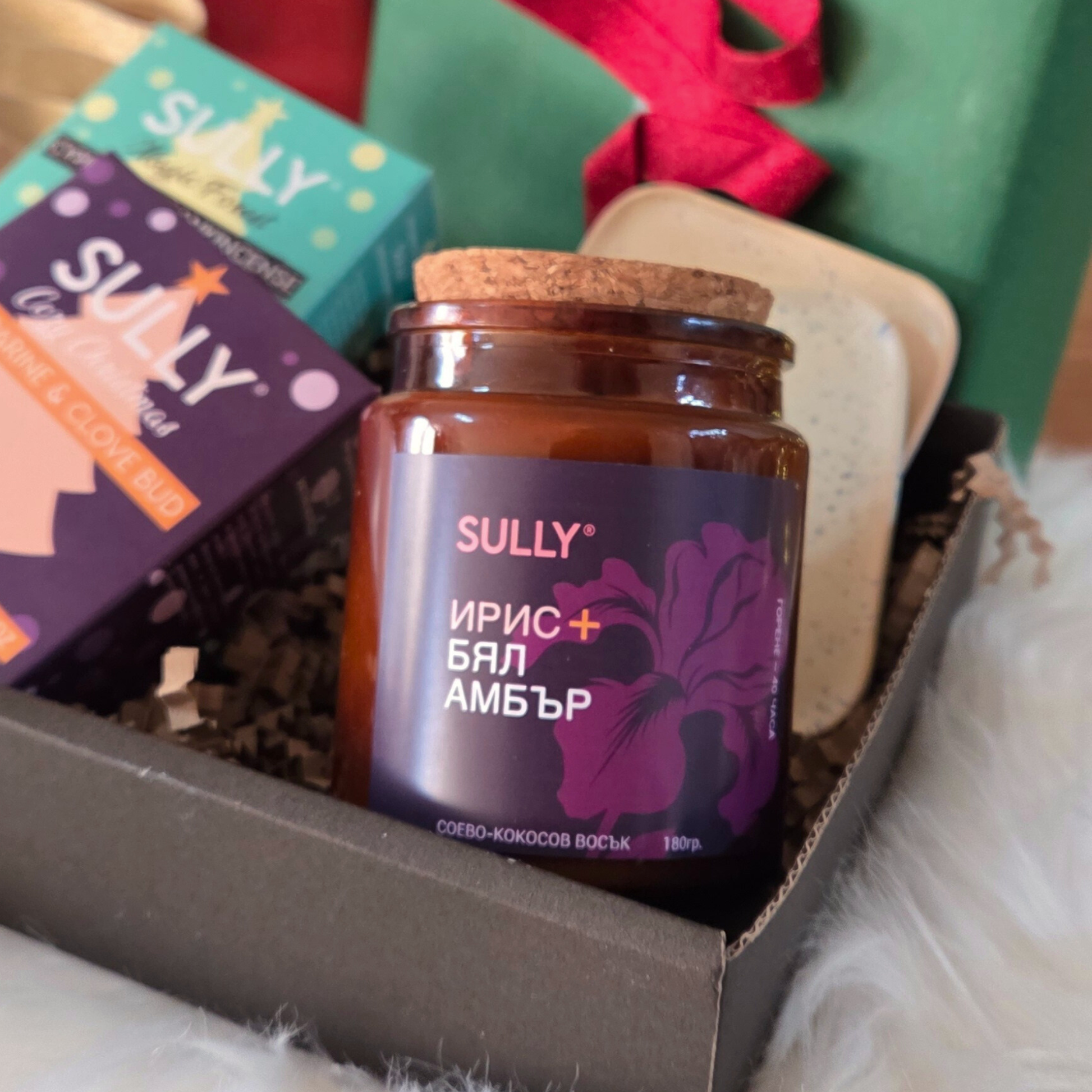 Свещ „Ирис и бял амбър“ от комплекта „Зимна грижа“ SULLY Naturals, заснета сред празнична украса и сапуни.
